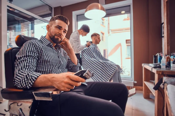 Homem elegante jovem na camisa quadriculada está sentado na barbearia ocupada e segurando aparador para corte de cabelo — Fotografia de Stock