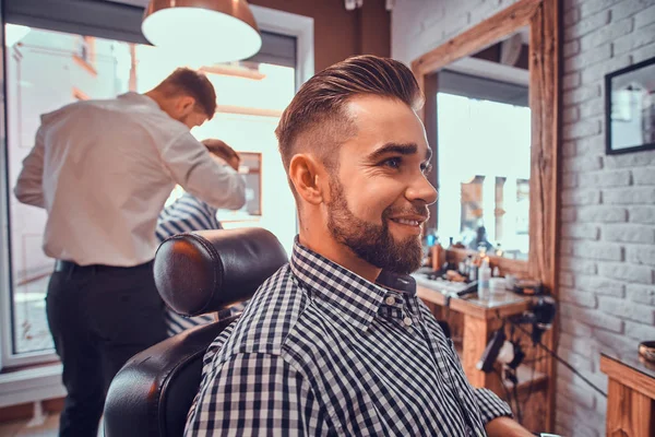 Atraente homem preparado está sentado enquanto espera por um barbeiro na barbearia ocupada — Fotografia de Stock