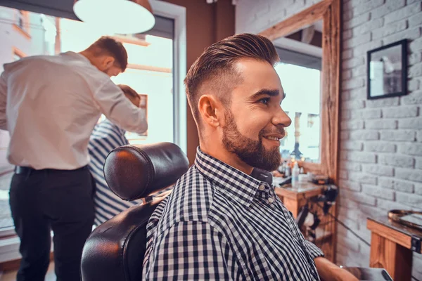 Atraente homem preparado está sentado enquanto espera por um barbeiro na barbearia ocupada — Fotografia de Stock