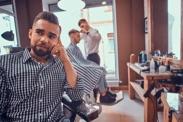 Atraente homem preparado está sentado enquanto espera por um barbeiro na barbearia ocupada — Fotografia de Stock