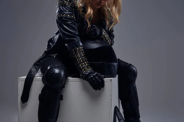 Fashion, sport, extreme. Cropped image of a biker girl wearing motorcycle gear sitting on a gray box in a studio. — Stock Photo, Image