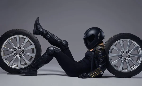 Moda, desporto, extremo. Menina motociclista vestindo equipamento de motocicleta deitado em um chão com rodas de carro em um estúdio . — Fotografia de Stock