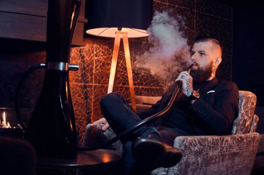 Pensive dövmeli adam koltukta oturuyor ve duman nargile başlıyor