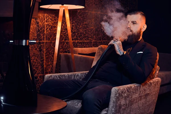 Nachdenklich tätowierter Mann sitzt auf dem Sessel und beginnt Wasserpfeife zu rauchen — Stockfoto