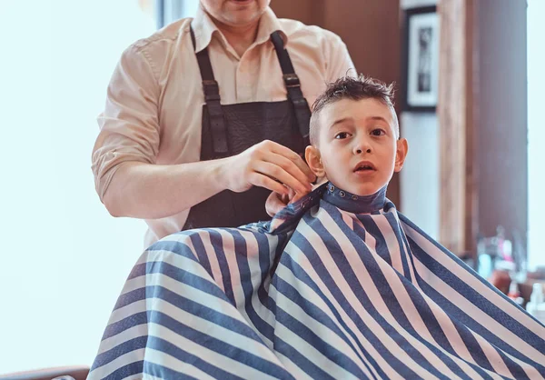 Little charming boy is getting trendy haircut from mature hairdresser at fashionable hairdressing salon — Stock Photo, Image