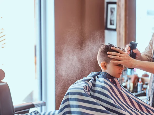 Little charming boy is getting trendy haircut from mature hairdresser at fashionable hairdressing salon — Stock Photo, Image