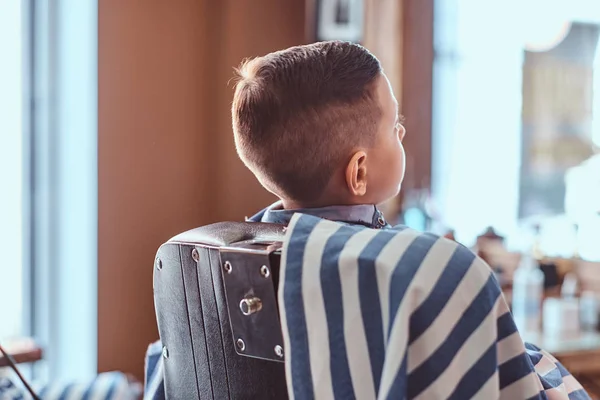 Pequeno menino bonito está mostrando seu novo penteado enquanto sentado na barbearia . — Fotografia de Stock