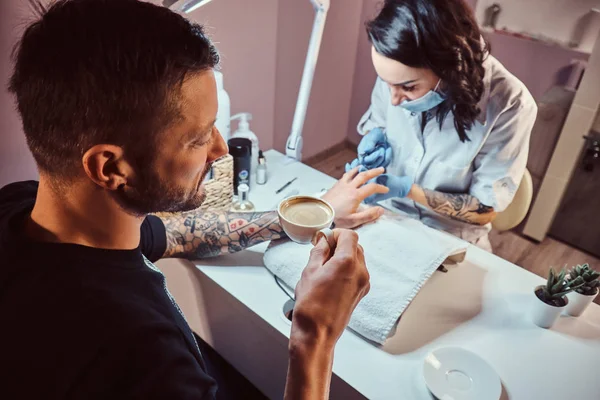 A man receiving a manicure in the beauty salon, relaxing and drinking a coffee while beautician master does manicure procedure — Stock Photo, Image