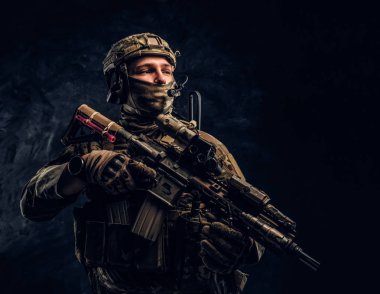 Fully equipped soldier in camouflage uniform holding an assault rifle. Studio photo against a dark wall clipart