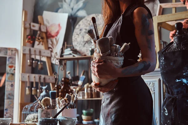 Giovane attraente pittore è holdind vaso con pennelli nel suo laboratorio accogliente — Foto Stock