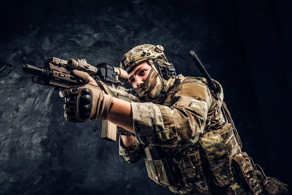 La unidad de élite, soldado de las fuerzas especiales en uniforme de camuflaje que sostiene un rifle de asalto con una mira láser y apunta al objetivo. Foto del estudio contra una pared oscura — Foto de Stock