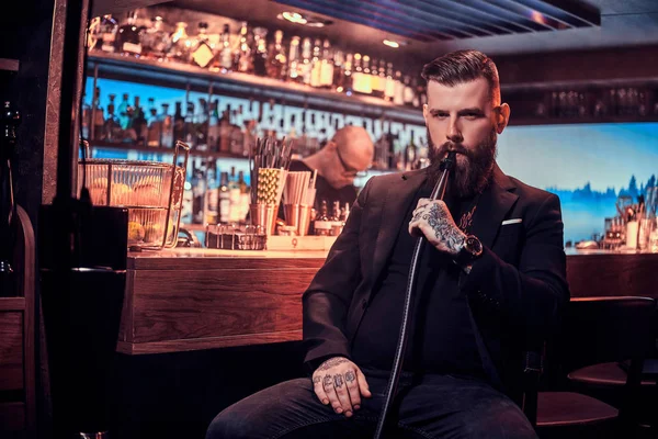 Atractivo hombre arreglado con barba y tatuajes está fumando narguile mientras está sentado cerca del mostrador del bar —  Fotos de Stock