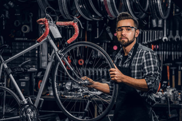 Attractive pensive man in protective glasses is repairing bicycle at his own workshop