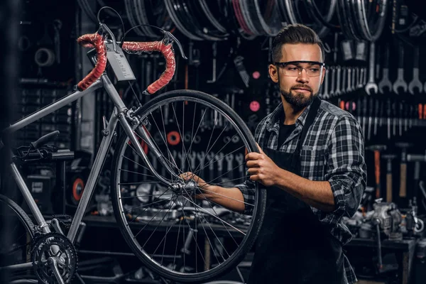 Attractive pensive man in protective glasses is repairing bicycle at his own workshop