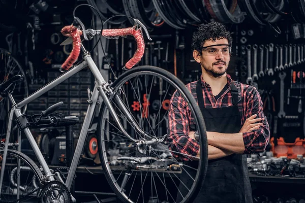 Smiling man in glasses is standing near fixed bicycle at his own workshop.