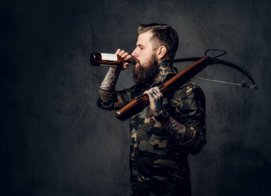 Askeri gömlekli sakallı dövmeli hipster adam elinde ortaçağ tatar yayı tutuyor ve bir zanaat birası içiyor. Karanlık duvara karşı stüdyo fotoğrafı