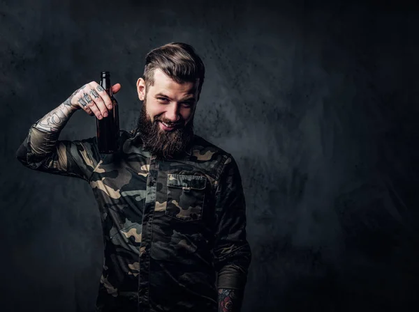 Elegante tipo hipster barbudo con camisa militar sosteniendo una cerveza artesanal y mirando una cámara. Estudio foto contra la pared oscura —  Fotos de Stock
