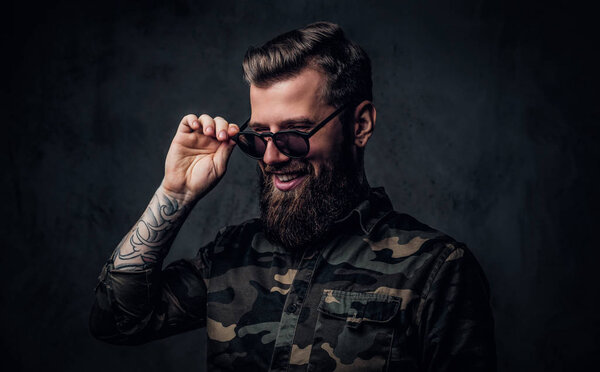 Portrait of a stylish bearded guy with tattooed hands in the military shirt holding hand on sunglasses and laughs. Studio photo against dark wall