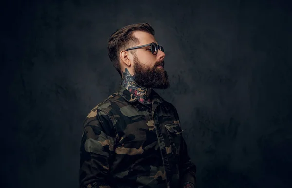 Portrait d'un homme barbu élégant avec les mains tatouées dans la chemise militaire. Studio photo contre mur sombre — Photo