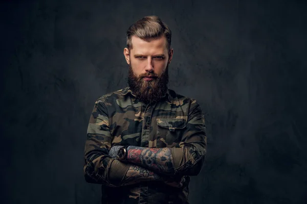 Ritratto di un ragazzo barbuto elegante con le mani tatuate nella camicia militare. Studio foto contro muro scuro — Foto Stock