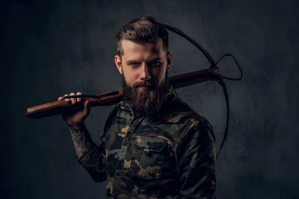 Un tipo hipster tatuado barbudo con camisa militar posando con una ballesta medieval. Estudio foto contra la pared oscura — Foto de Stock