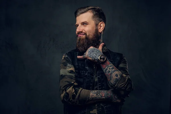 Un tipo barbudo divertido con manos tatuadas mostrando un signo de shaka y mirando una cámara. Foto del estudio contra una pared oscura — Foto de Stock