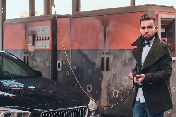 Hombre atractivo está posando con la manguera en la estación de lavado de coches cerca de su coche . — Foto de Stock