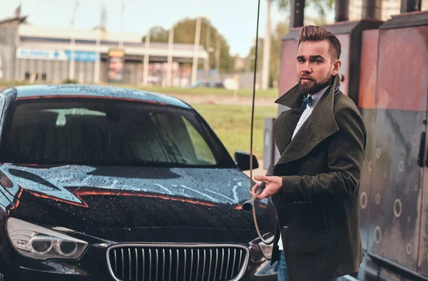 Hombre atractivo está posando con la manguera en la estación de lavado de coches cerca de su coche . — Foto de Stock