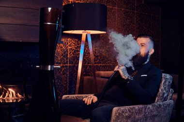 Pensive dövmeli adam koltukta oturuyor ve duman nargile başlıyor