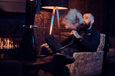 Pensive dövmeli adam koltukta oturuyor ve duman nargile başlıyor