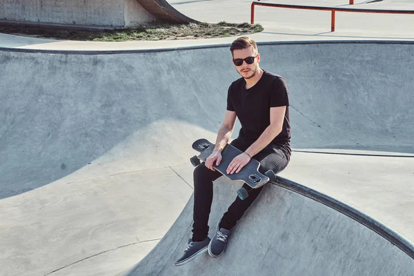 Joven patinador en gafas de sol está sentado en la rampa con su propio longboard . — Foto de Stock