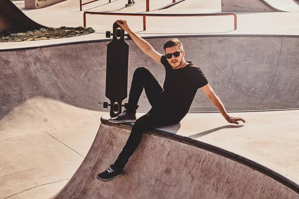 Joven patinador en gafas de sol está sentado en la rampa con su propio longboard . — Foto de Stock