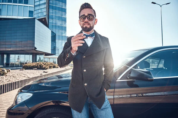 Hombre guapo en abrigo está de pie cerca de su nuevo coche mientras fuma en el aparcamiento . — Foto de Stock