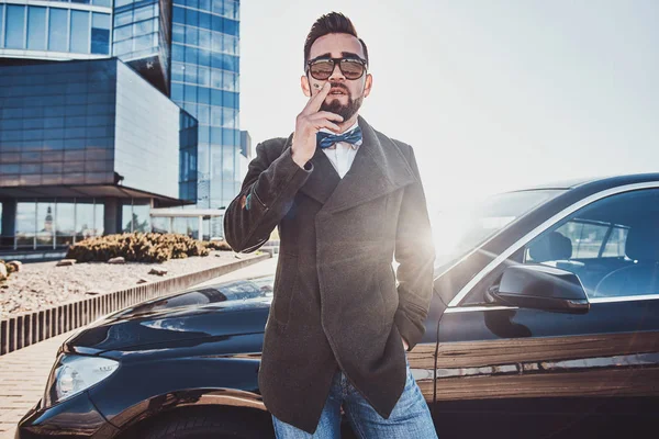 Hombre guapo en abrigo está de pie cerca de su nuevo coche mientras fuma en el aparcamiento . — Foto de Stock