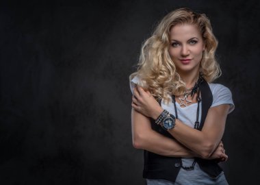 Beyaz bir t-shirt ve yelek giymiş Şehvetli kıvırcık sarışın kadın bir stüdyoda çapraz kolları ile poz, aksesuar ve kol saati bir sürü giyer. Koyu dokulu arka planda yalıtılmış.