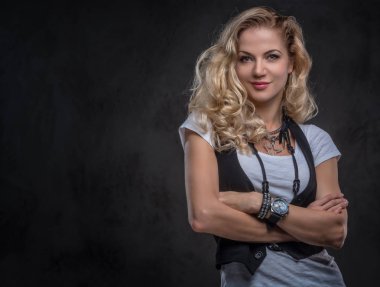 Beyaz bir t-shirt ve yelek giymiş Şehvetli kıvırcık sarışın kadın bir stüdyoda çapraz kolları ile poz, aksesuar ve kol saati bir sürü giyer. Koyu dokulu arka planda yalıtılmış.