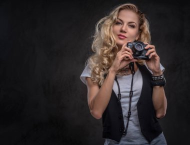 Beyaz bir t-shirt ve yelek giymiş Şehvetli kıvırcık sarışın kız fotoğrafçı aksesuar ve kol saati bir sürü giyer, bir stüdyoda bir kamera ile poz. Koyu dokulu arka plan da yalıtılmış.