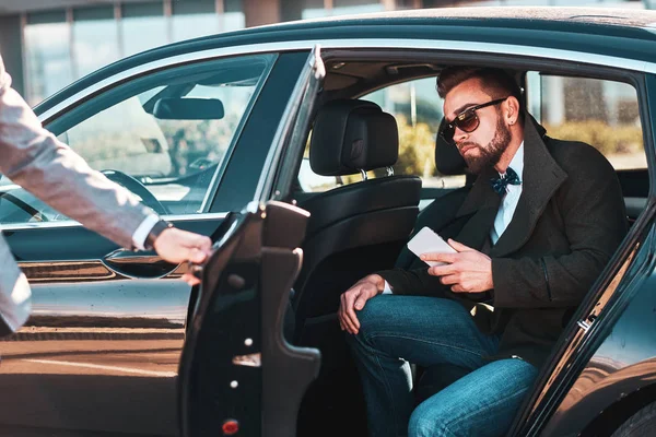 Απασχολημένος καλός επιχειρηματίας με γυαλιά ηλίου μιλάει με το smartphone, ενώ κομψή βοηθό του ανοίγει την πόρτα για αυτόν. — Φωτογραφία Αρχείου