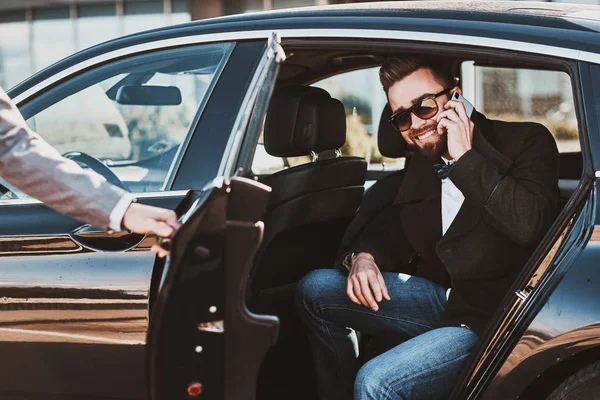 Ocupado agradable hombre de negocios en gafas de sol está hablando por teléfono inteligente, mientras que su elegante asistente está abriendo la puerta para él . — Foto de Stock