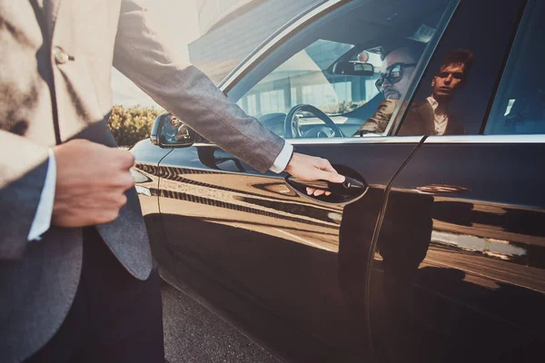 Empresário inteligente está sentado no carro enquanto seu jovem assistente está abrindo a porta para ele . — Fotografia de Stock