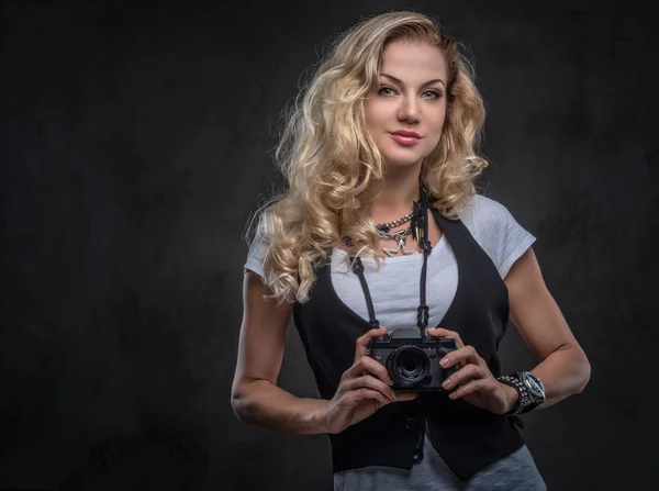 Sensuelle fille blonde bouclée photographe vêtue d'un t-shirt blanc et gilet porte beaucoup d'accessoires et de montre-bracelet, posant avec un appareil photo dans un studio. Isolé sur un fond texturé foncé . — Photo