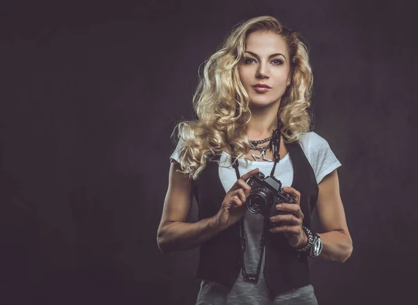 Sensuele krullend blonde meisje fotograaf gekleed in een wit t-shirt en gilet draagt veel accessoires en polshorloge, poseren met een camera in een studio. Geïsoleerd op een donkere getextureerde achtergrond. — Stockfoto