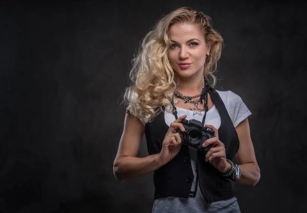 Sensuele krullend blonde meisje fotograaf gekleed in een wit t-shirt en gilet draagt veel accessoires en polshorloge, poseren met een camera in een studio. Geïsoleerd op een donkere getextureerde achtergrond. — Stockfoto