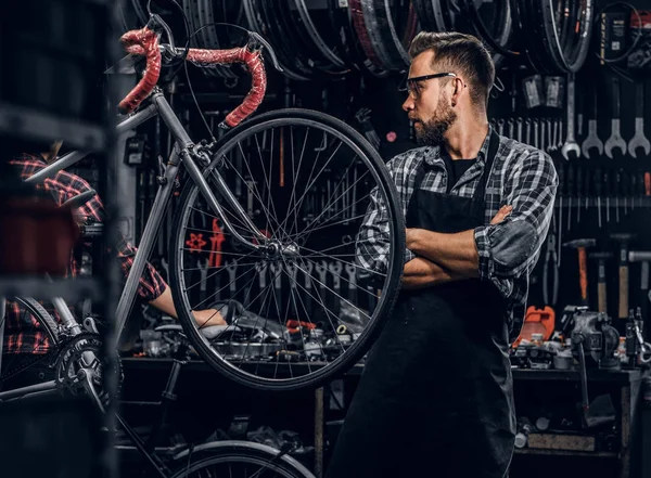 Красивый бородатый мужчина в очках стоит возле неподвижного велосипеда в своей мастерской — стоковое фото