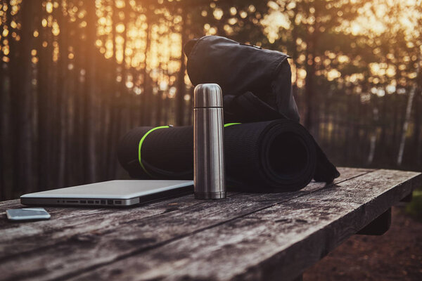 Silver laptop, smartphone , black backpack, mat and thermos on the wooden table in the forest.