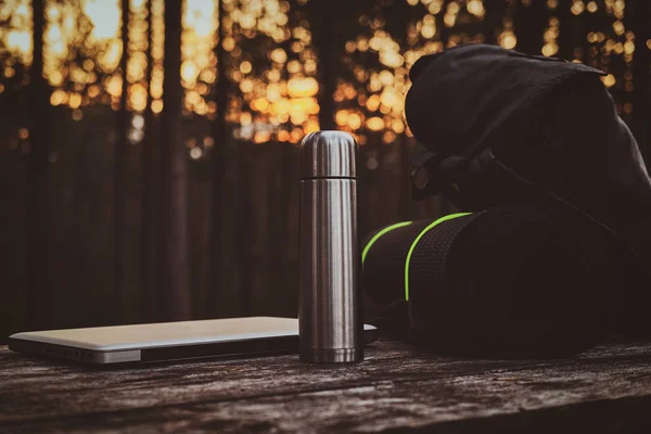 Portátil de plata, smartphone, mochila negra, estera y termo en la mesa de madera en el bosque . — Foto de Stock