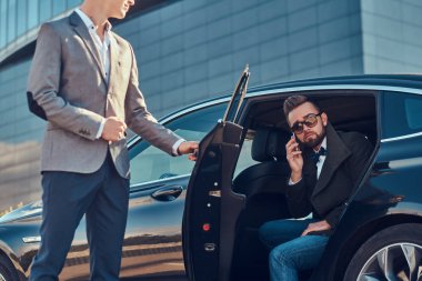 Güneş gözlüğü çekici bakımlı adam akıllı telefon tarafından konuşuyor ve asistanı onun için kapıyı açarken arabada oturuyor.
