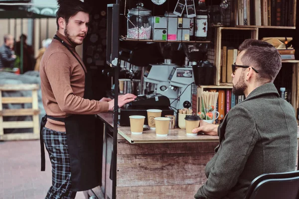 El hombre elegante arreglado en gafas de sol está bebiendo café y hablando con barista mientras está sentado afuera en coffeeshop — Foto de Stock