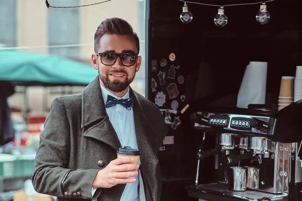 El hombre sonriente moderno en gafas de sol está tomando café mientras está parado afuera en coffeeshop — Foto de Stock