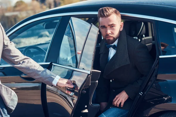 Empresário atraente inteligente está saindo do carro enquanto seu assistente está abrindo a porta para ele . — Fotografia de Stock